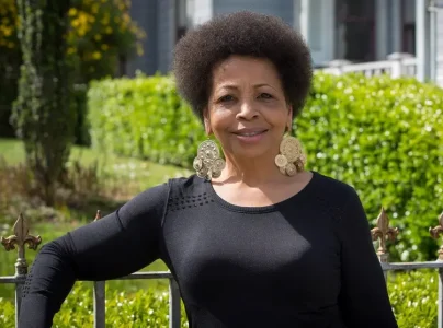A person with short, curly hair and large circular earrings stands outdoors, leaning on a wrought iron fence with greenery and an SFPD commission building in the background.