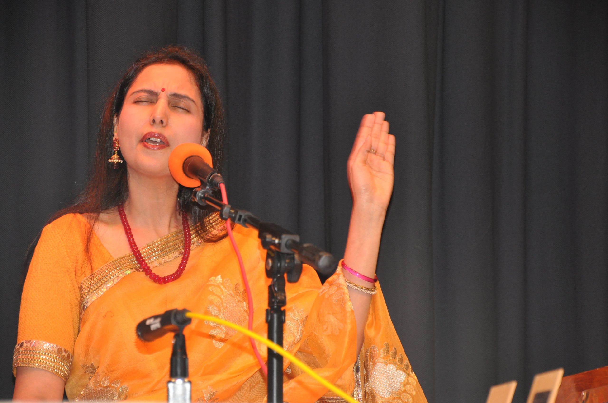 A woman in a sari sings into a microphone with eyes closed and one hand raised, in front of a dark curtain.