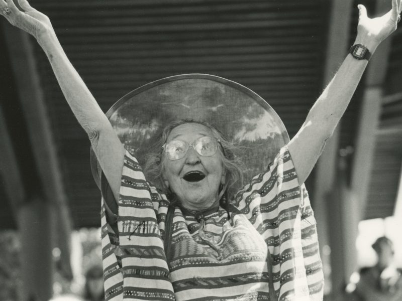 An elderly woman with glasses and a striped dress raises her arms joyfully, wearing a large, round hat. The background shows a partially visible structure.