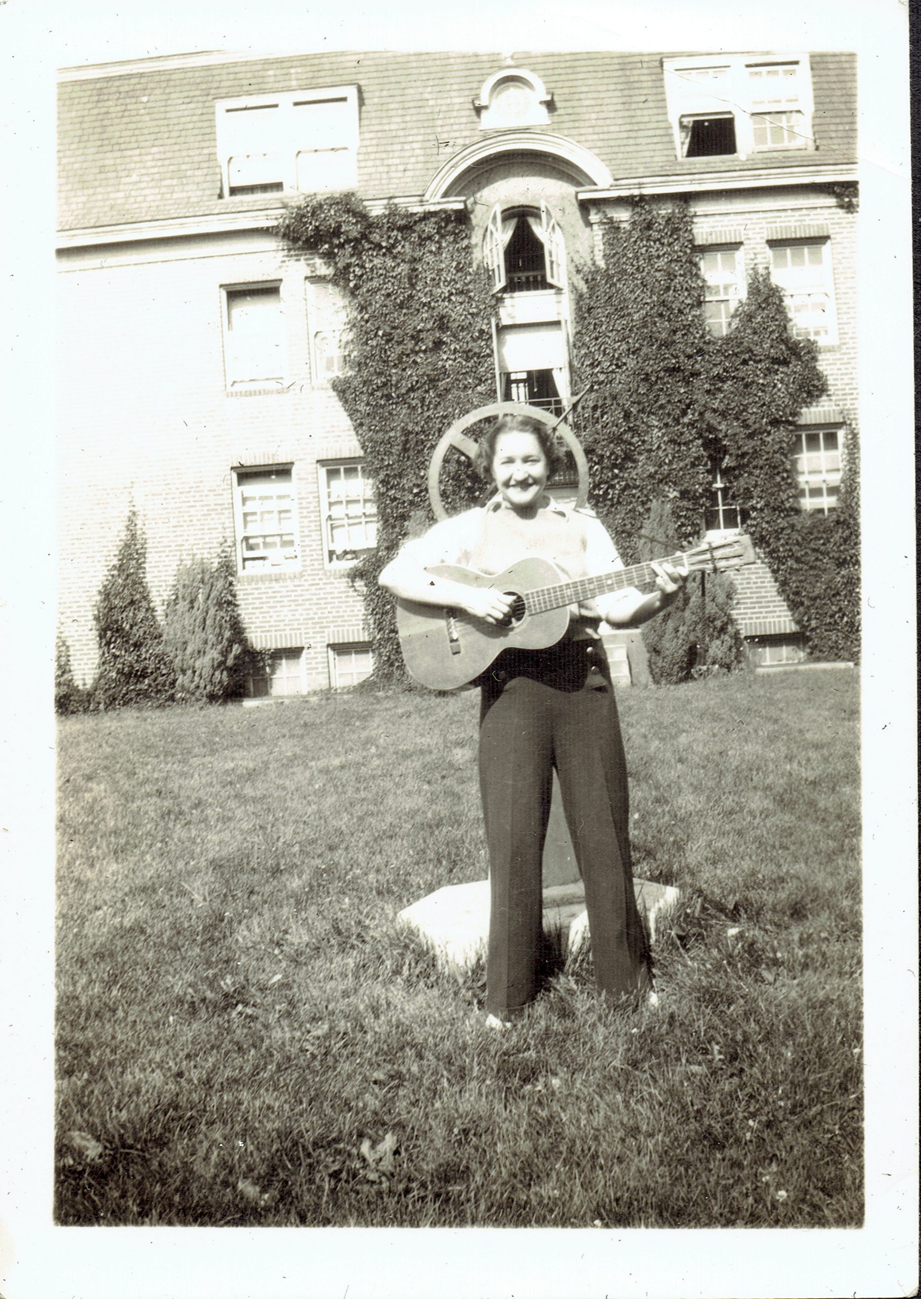 Person standing on grass, playing a guitar in front of a building covered partially in ivy.