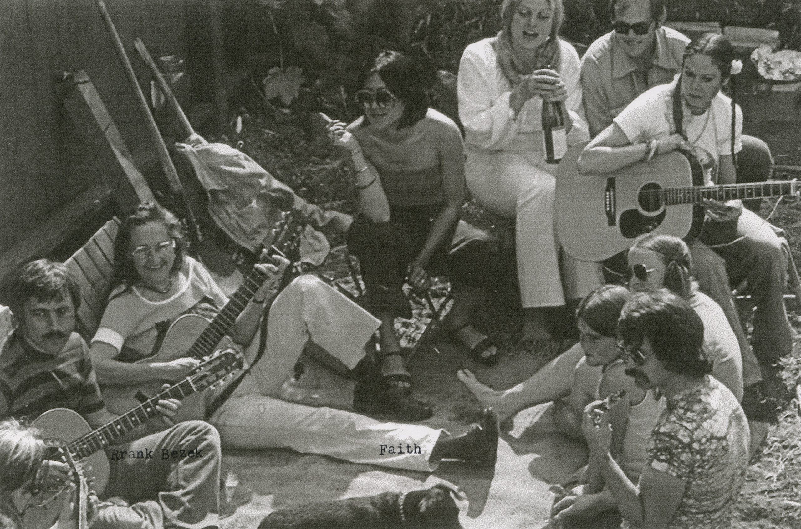 A group of people sit on the grass enjoying a casual gathering. Two individuals play acoustic guitars while others relax and chat, with drinks and a dog nearby.