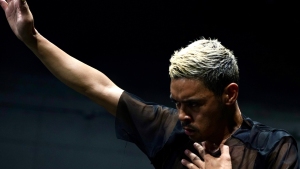 A person with short, bleached hair wearing a sheer black top raises one arm while placing the other hand on their chest against a dark background.