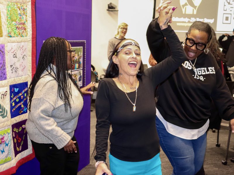 Three people are joyfully dancing near a colorful quilt. A woman in a black shirt and blue pants raises her arms, smiling broadly as others join in the celebration.
