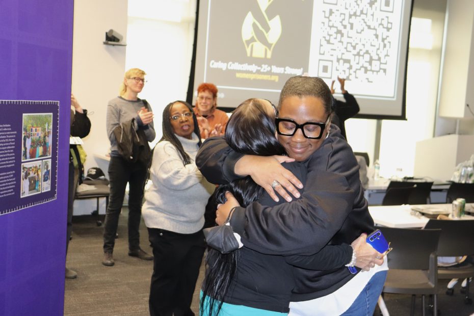 Two people hugging in a room with a small audience. A presentation slide with a QR code is displayed in the background.