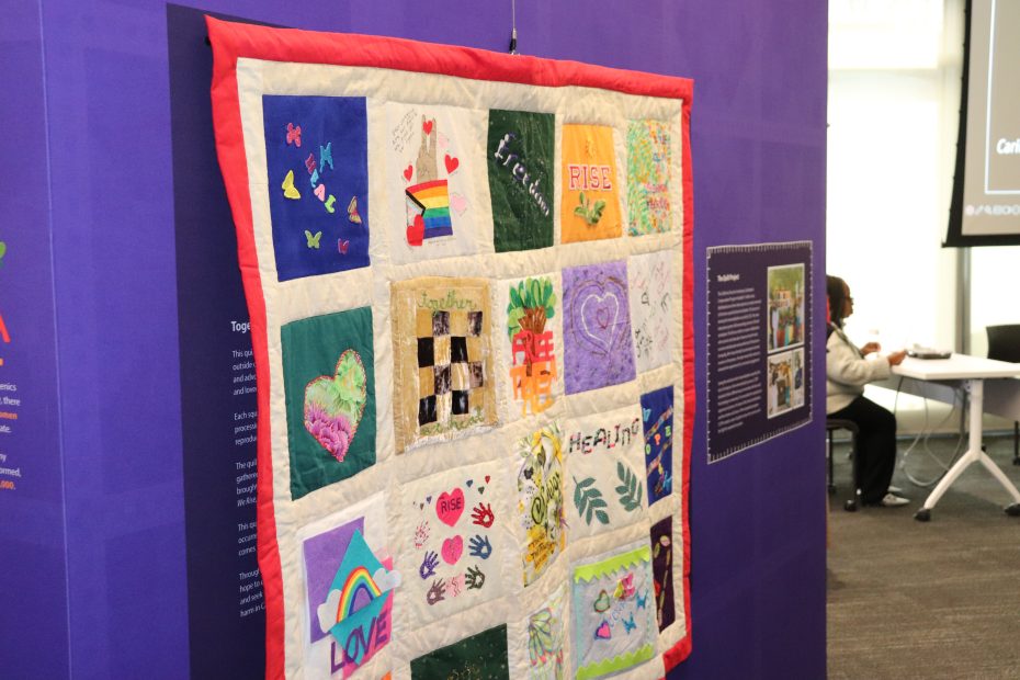 Quilt with colorful squares featuring various designs and words, hung on a purple wall display.