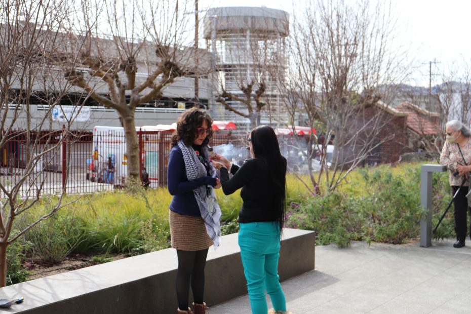 Two women are standing outside near a planter. One is handing an object to the other. Leafless trees and a building are in the background.