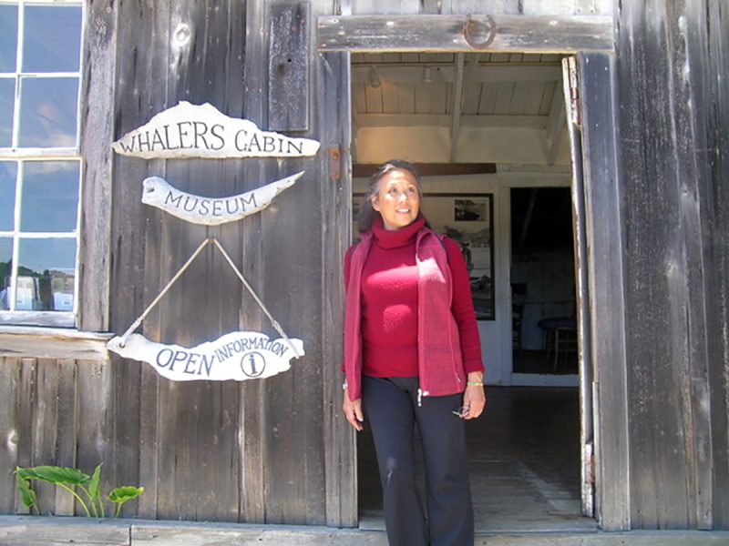 A person stands at the entrance of Whalers Cabin Museum, beside signs labeled "Whalers Cabin" and "Open Information." The cabin is wooden with a rustic appearance.