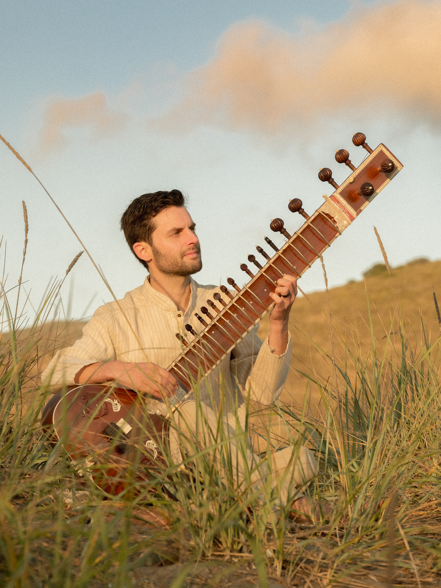 Person sitting outdoors in grass, playing a sitar, with a cloudy sky and hills in the background.