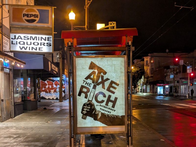 Bus stop poster at night featuring an advertisement with the slogan "Axe the Rich" and a hand holding an axe. Street lights and stores are visible in the background.