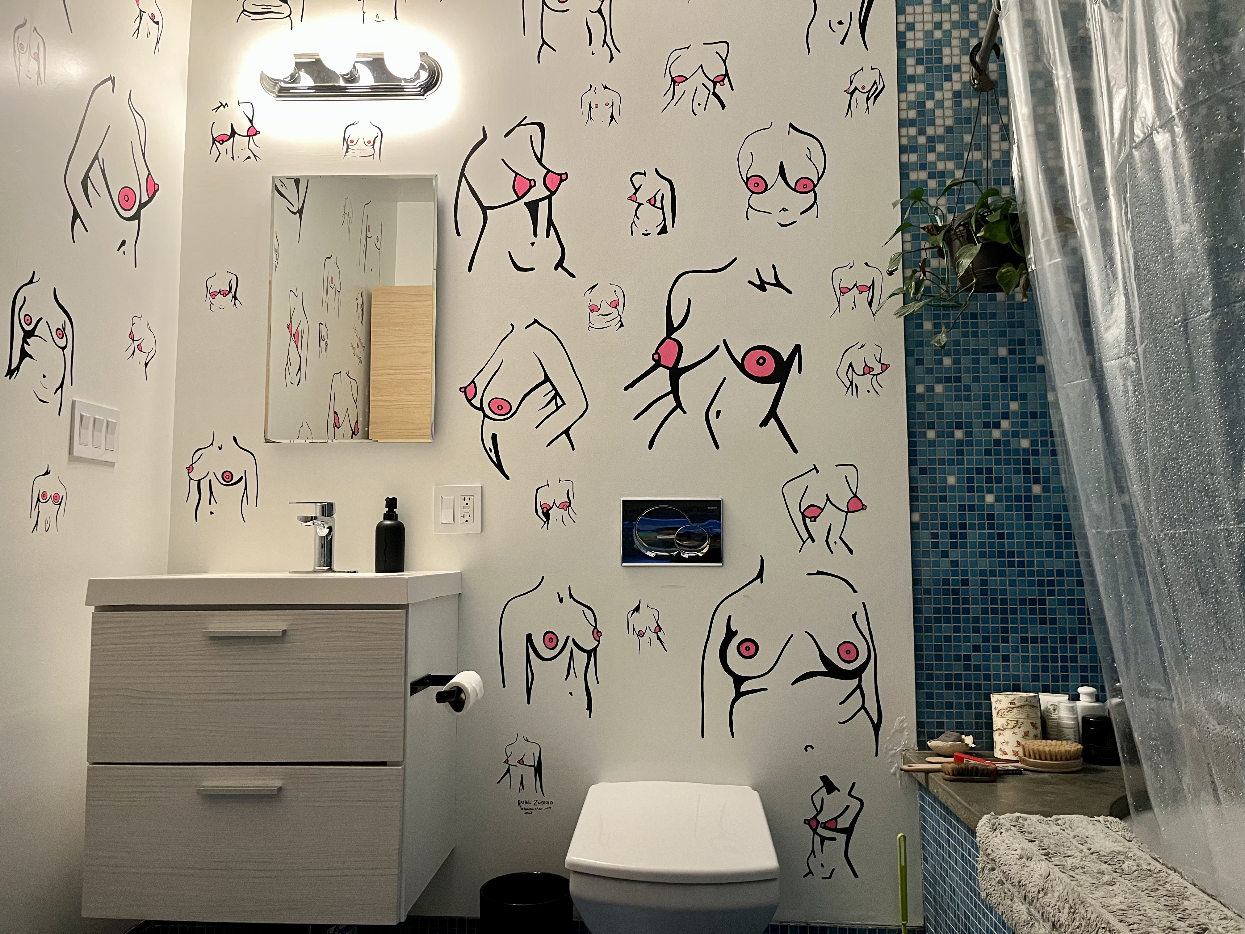Bathroom with white walls featuring abstract face illustrations in black with pink accents. Blue tiled area near shower. White sink and toilet, with various toiletries around.