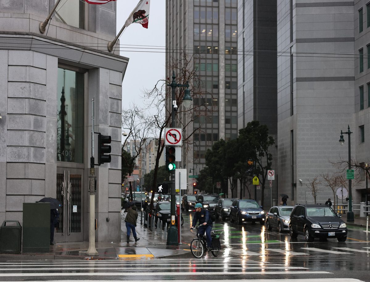 Rainy city street with cars stopped at a traffic light and pedestrians crossing the road. Umbrellas and wet pavement are visible. Tall buildings line the street.