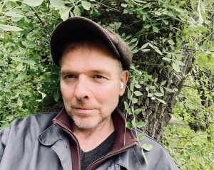 Person wearing a cap and earphones stands outdoors near a tree with green leaves.