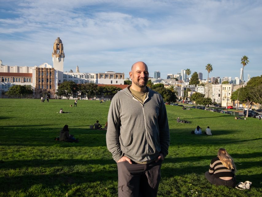 A person stands smiling in a green park with a cityscape and clock tower in the background.