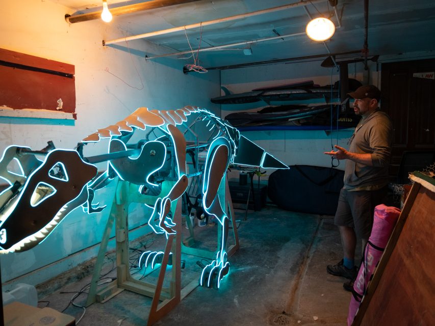 A man observes a glowing dinosaur skeleton model in a dimly lit garage.