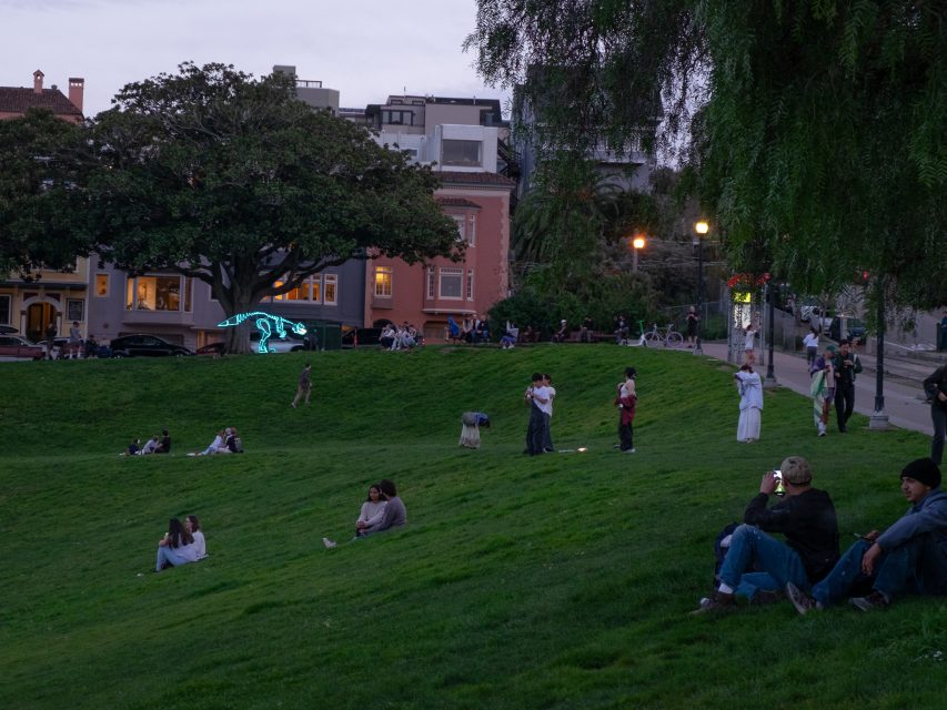People sit and walk on a grassy slope in a park during the evening. Trees and buildings are visible in the background.