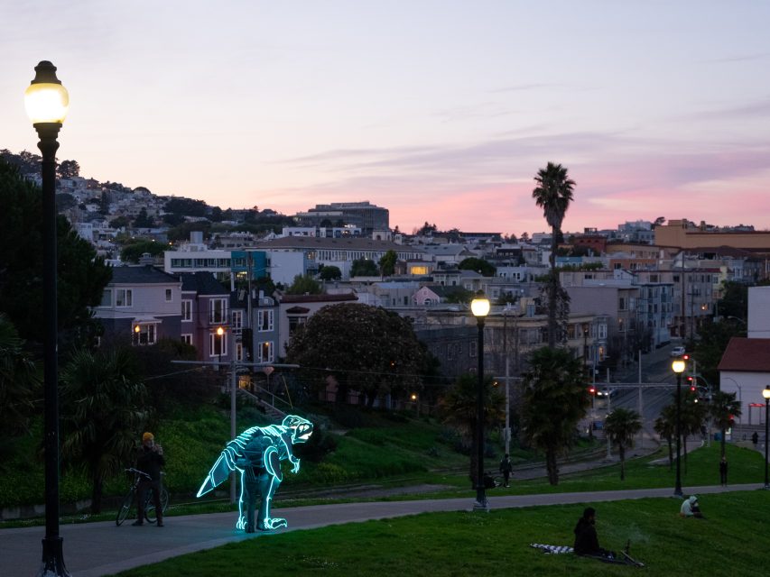 A glowing neon dinosaur sculpture in a park during dusk, with people sitting on the grass and a cityscape in the background.