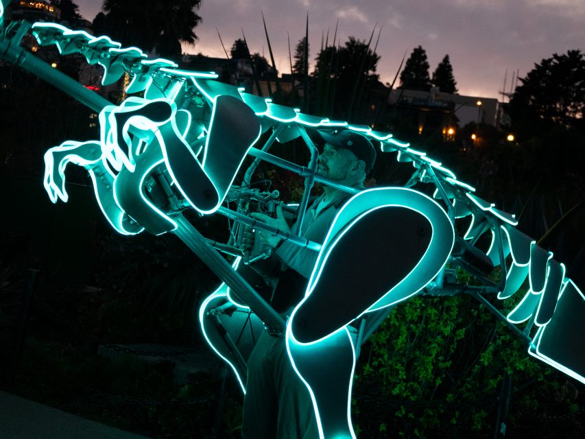 Person wearing a glowing dinosaur skeleton costume, illuminated in blue, stands outdoors at dusk with trees and buildings in the background.