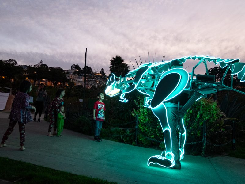 Children observe a person wearing a glowing dinosaur costume outdoors at dusk.