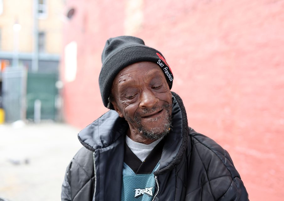 An elderly man in a black beanie and jacket stands smiling with eyes closed, against a red brick wall and blurred urban background, while the city quietly conducts its own triage.