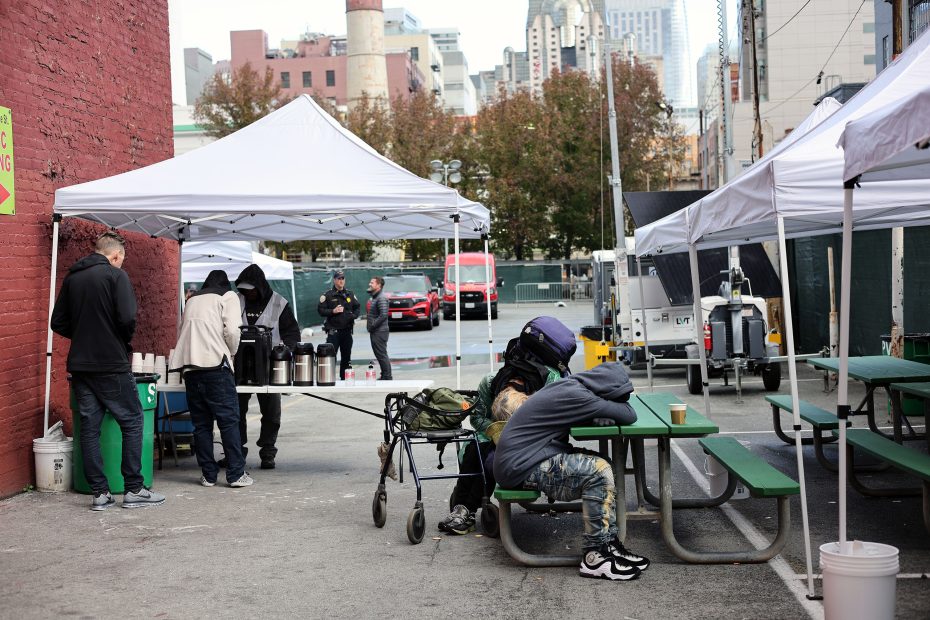 People gather at outdoor tents with tables, one person resting on a bench. With city buildings visible in the background, the scene resembles a casual triage setup amid an urban landscape.