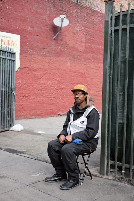 A person in a jacket and hat sits on a small chair, overseeing triage efforts on the sidewalk near a gate. A red brick wall with a satellite dish looms in the background, adding an urban touch to the scene.
