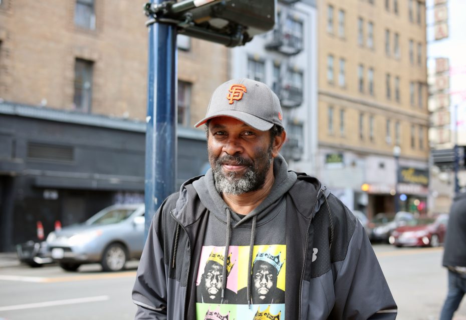 A man wearing a San Francisco Giants cap and graphic hoodie stands on a bustling city street, blending into the urban triage of buildings and cars around him.