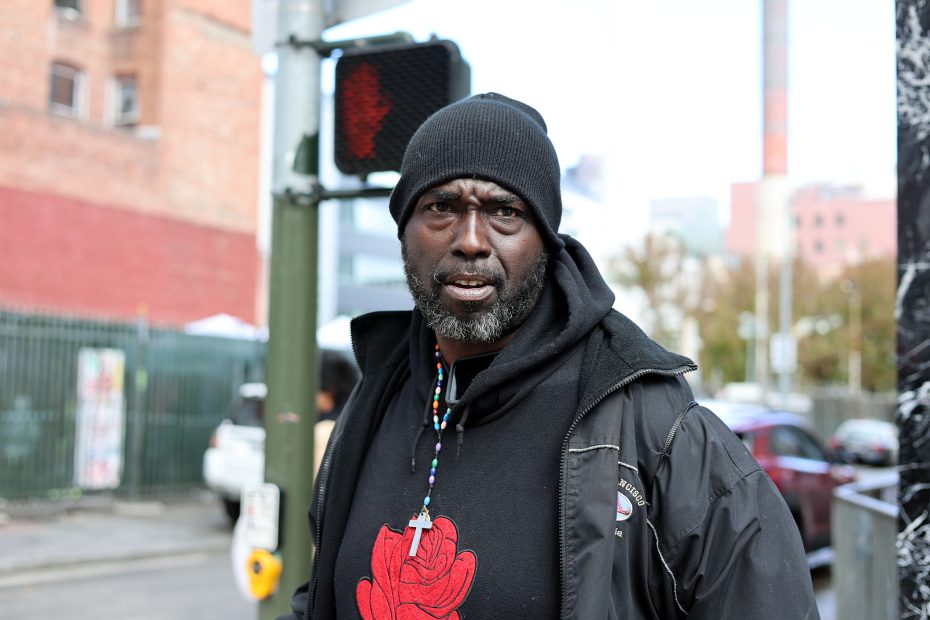 A man in a black beanie and jacket stands on a bustling city street, the red pedestrian signal in the background, as if waiting for his moment amidst the urban triage.