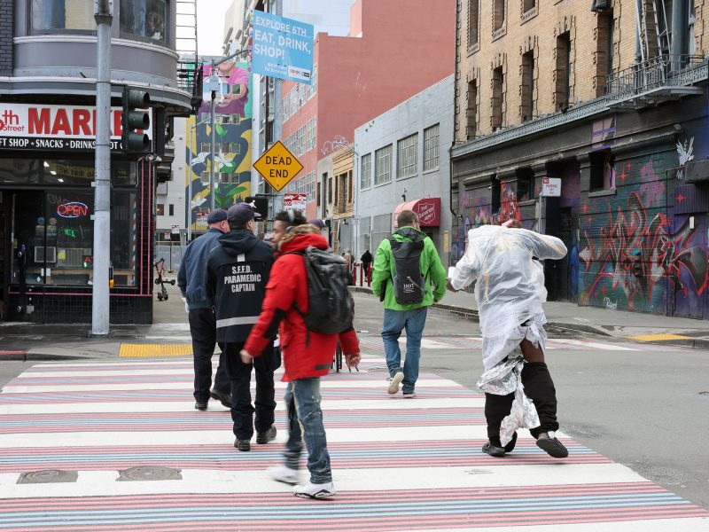 Amidst the vibrant cityscape, people cross the street, one draped in reflective material, reminiscent of a triage blanket. The backdrop of colorful buildings and dynamic street art adds an artistic flair to the urban scene.