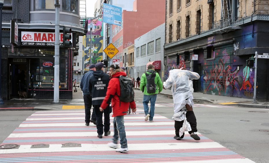 Amidst the vibrant cityscape, people cross the street, one draped in reflective material, reminiscent of a triage blanket. The backdrop of colorful buildings and dynamic street art adds an artistic flair to the urban scene.