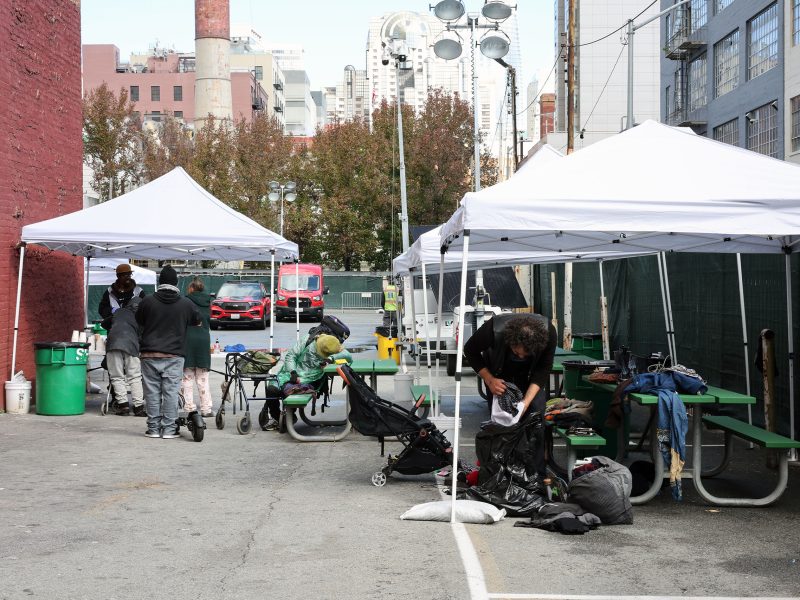 People gather under white tents set up in an urban alley, reminiscent of a triage area. Tables and personal belongings are visible, lending a sense of organized chaos. Buildings and trees provide a backdrop to this bustling scene.