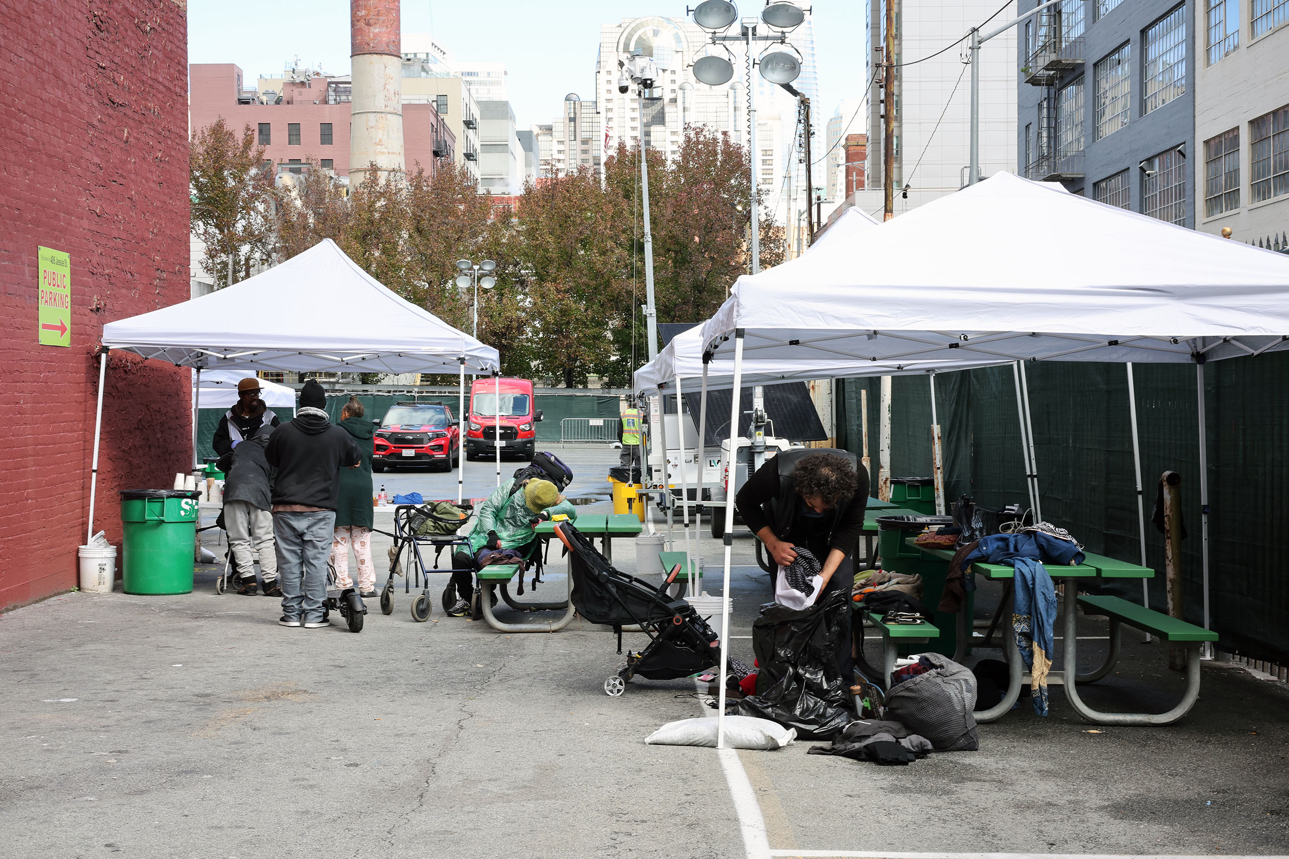 People gather under white tents set up in an urban alley, reminiscent of a triage area. Tables and personal belongings are visible, lending a sense of organized chaos. Buildings and trees provide a backdrop to this bustling scene.