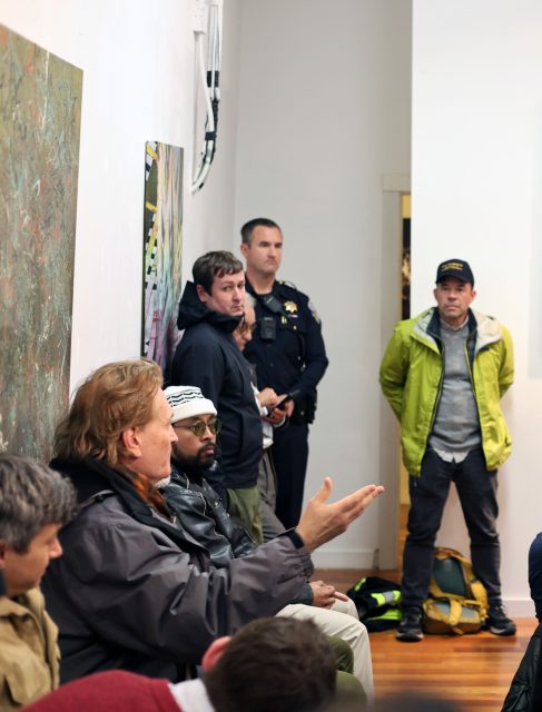 A group of people seated in a discussion, with one person gesturing. A police officer and others stand in the background. The setting appears to be an indoor space with art on the wall.