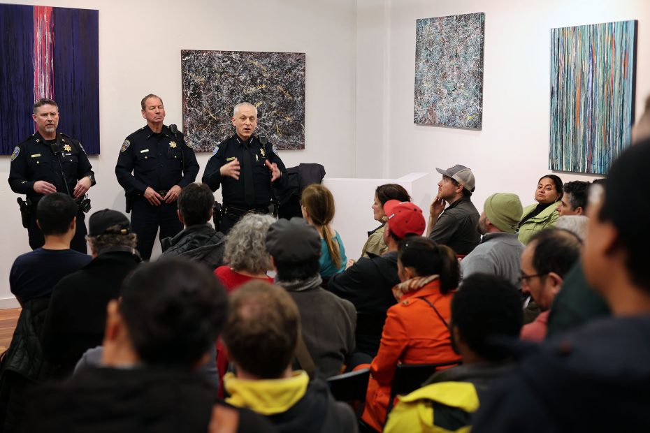 Police officers stand speaking to a seated audience in an art gallery, with abstract paintings on the walls.