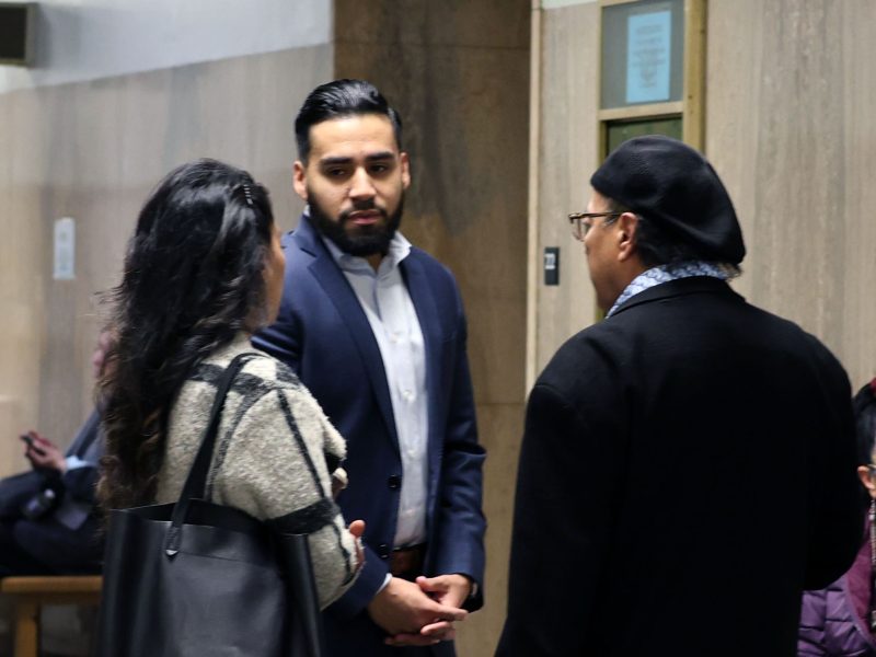 Three people conversing in a hallway: a woman with a handbag, a man in a suit, and another man in a beret and glasses. The background shows a wooden wall and a directory sign.