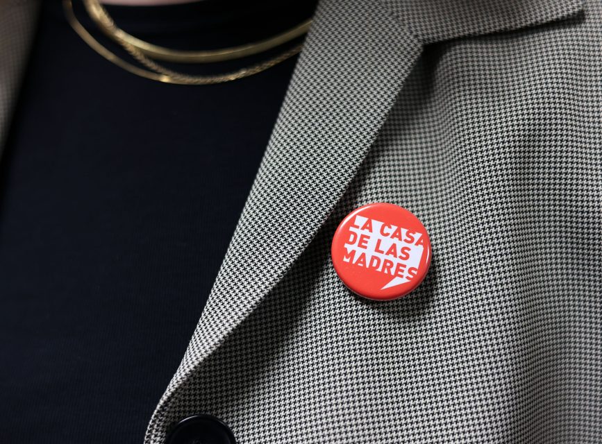 A person wearing a gray blazer and black shirt has a red button with the text "LA CASA DE LAS MADRES" pinned to the lapel.