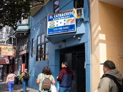 People walk past El Catrachito Restaurant on a city street. The restaurant's blue sign displays its name, a dish image, and operating hours.
