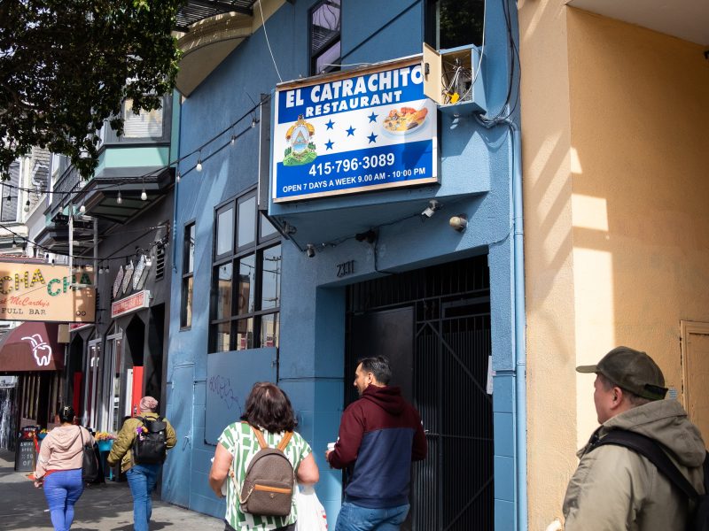 People walk past El Catrachito Restaurant on a city street. The restaurant's blue sign displays its name, a dish image, and operating hours.