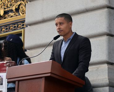 A person in a suit speaks at a podium with a microphone outside a building with ornate details.