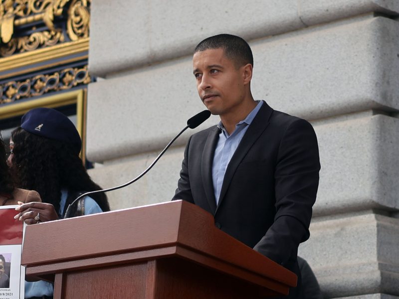 A person in a suit speaks at a podium with a microphone outside a building with ornate details.