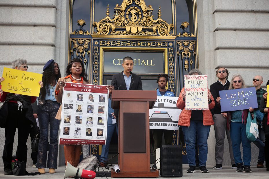 A speaker addresses a crowd outside City Hall, surrounded by people holding signs advocating for police reform and justice.