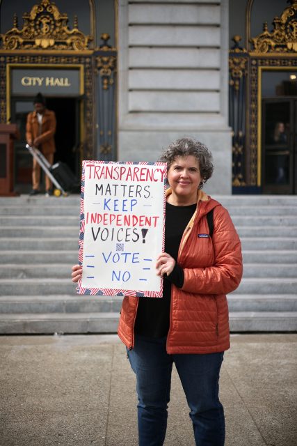 A person stands on city hall steps holding a sign that reads, "Transparency matters. Keep independent voices! Vote No," urging citizens to maintain integrity within the police commission.
