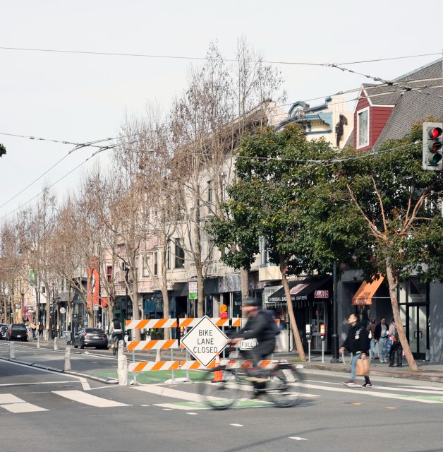 A cyclist rides past a "Bike Lane Closed" sign on an urban street with shops and bare trees.