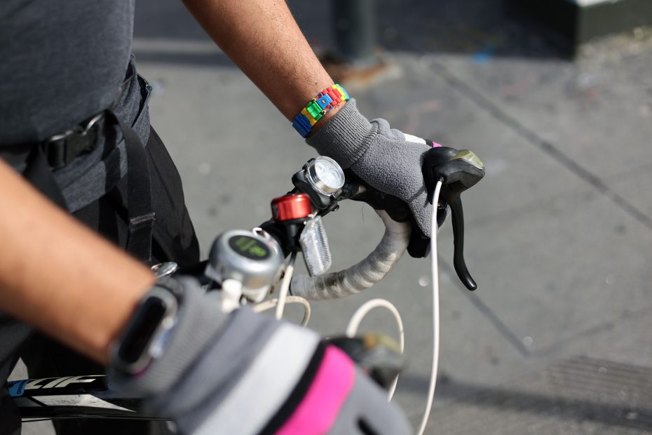 Person wearing gloves and colorful wristbands, holding the handlebars of a bicycle on a paved surface.