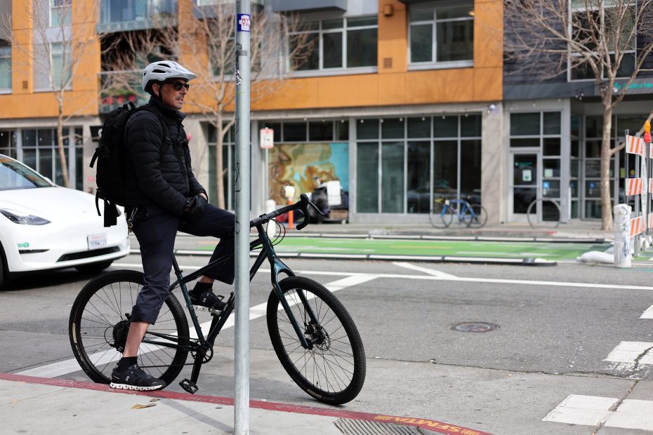 Person in cycling gear on a black bicycle waits at an intersection. A white car is parked nearby, and buildings are in the background.