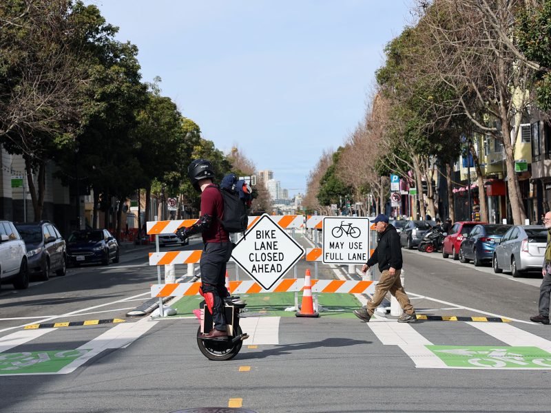 Person on a unicycle crosses a street with a "Bike Lane Closed" sign. Pedestrian crossing in the opposite direction. Cars and trees line the sides of the road.