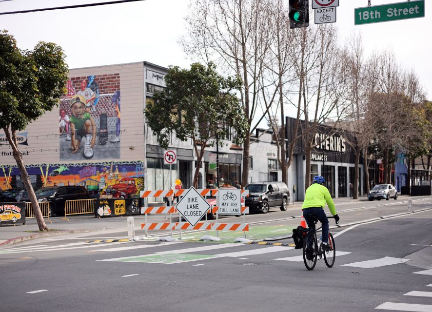 Cyclist in a green jacket rides past a "Bike Lane Closed" sign on a city street, with a mural and buildings visible in the background.