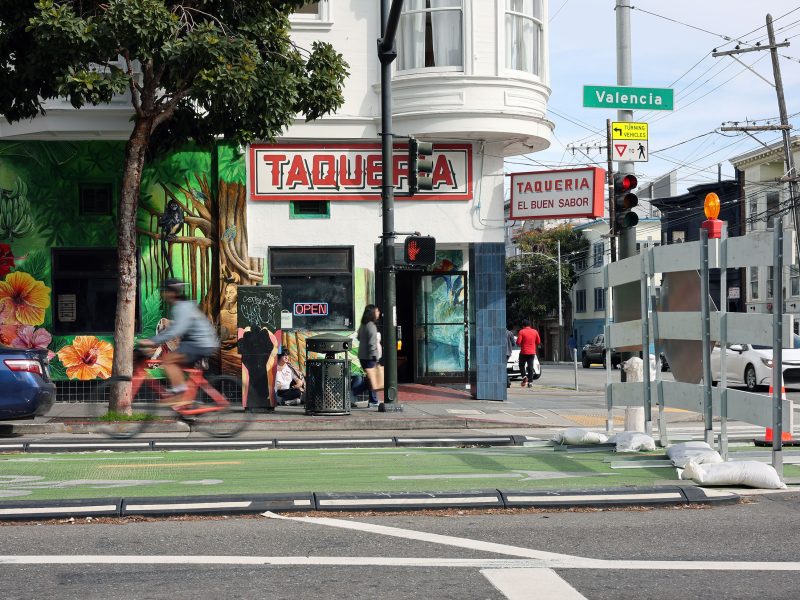 Street view of a taqueria with colorful mural. A cyclist rides past on a green bike lane, while pedestrians mill around. A sign for Valencia Street is visible. Traffic cones line the sidewalk.