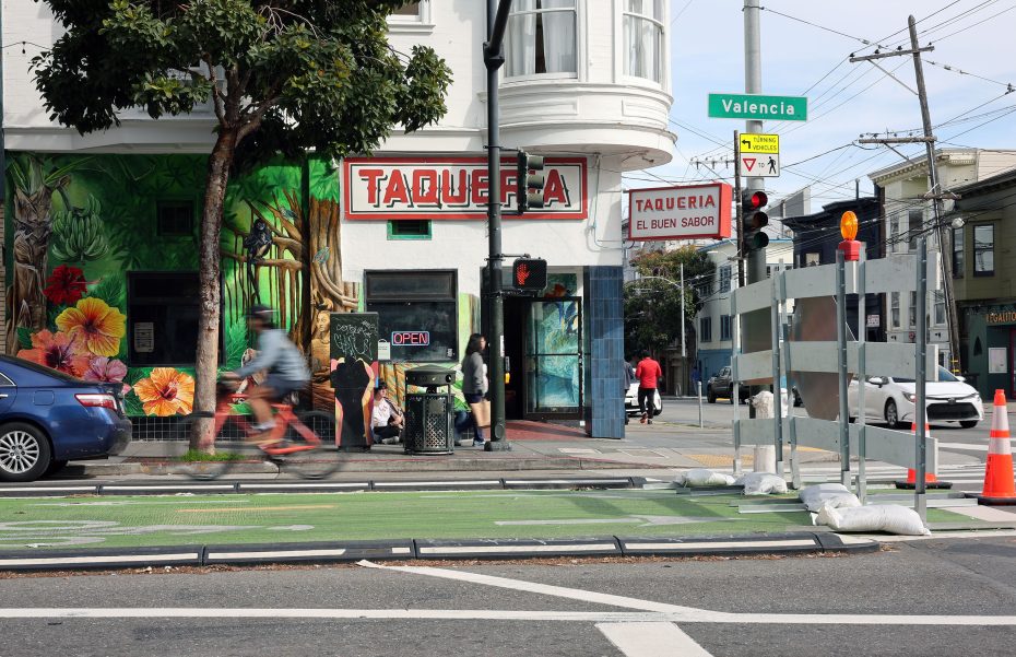 Street view of a taqueria with colorful mural. A cyclist rides past on a green bike lane, while pedestrians mill around. A sign for Valencia Street is visible. Traffic cones line the sidewalk.
