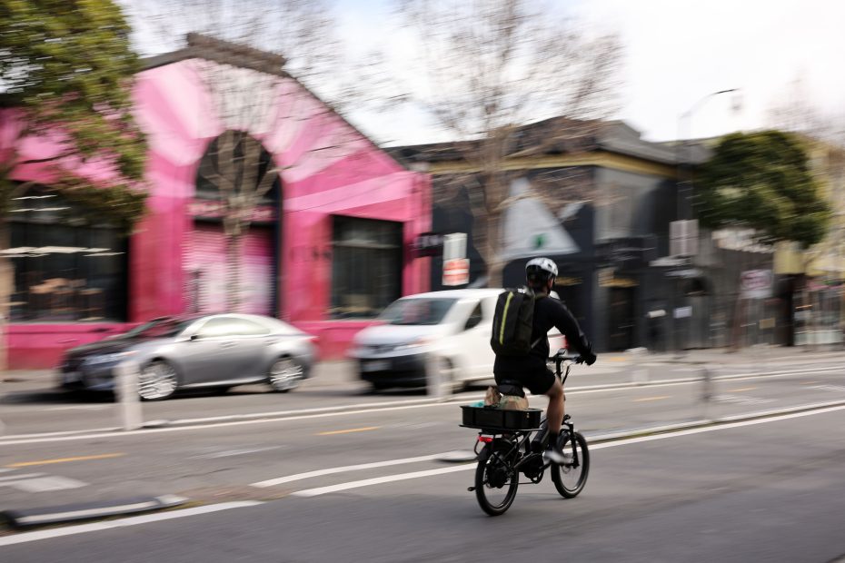 Person biking on a city street with colorful storefronts and cars in the background.