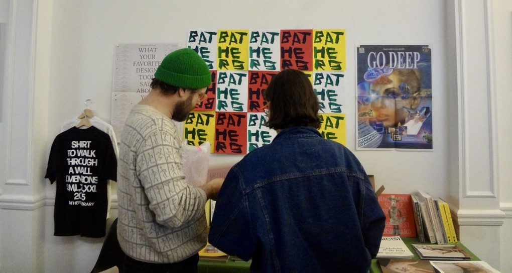 Two people stand at a table with books and magazines. Behind them are posters and a shirt on the wall.